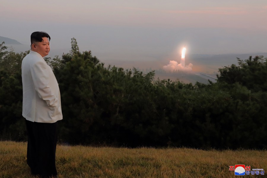 A landscape photo of an unknown place in North Korea. To the left in the foreground stands Kim Jong Un. To the right, in the background, a ballistic missile rises from its launch site.