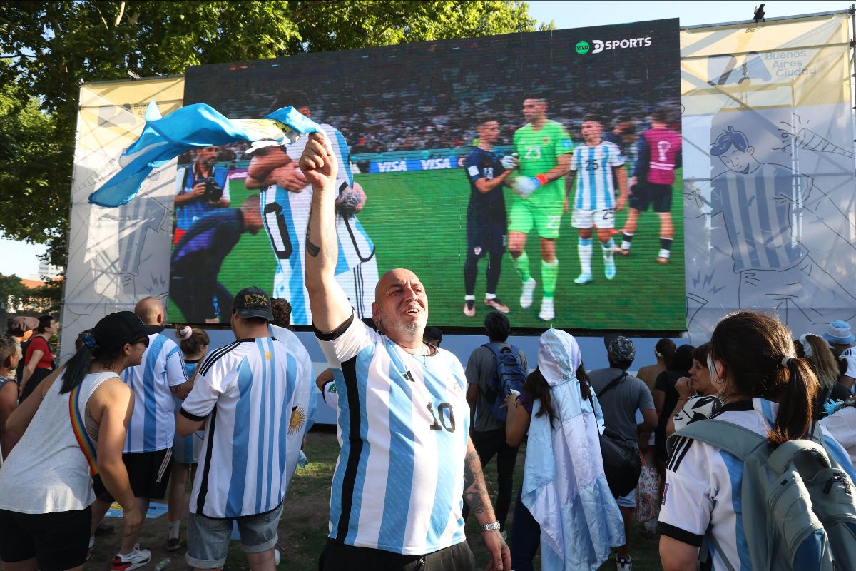Dozens of fans in an Argentina park are celebrating. Most of them are wearing jerseys of the national team. Behind them is a massive TV screen showing the players shaking hands with the referee.