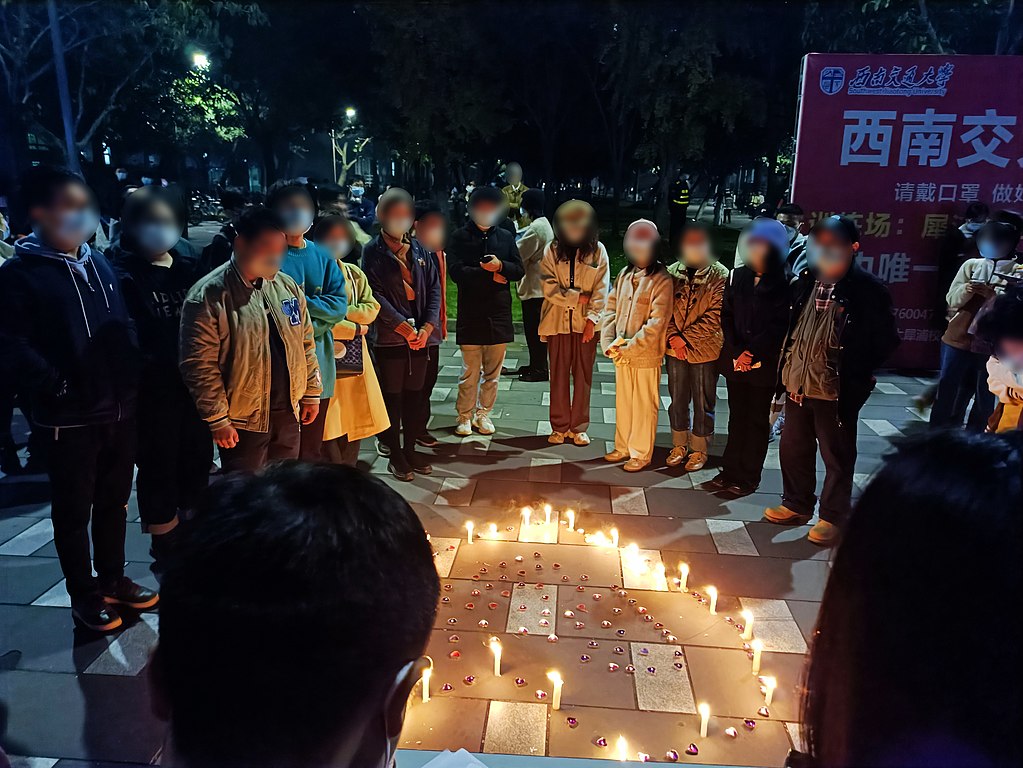 About a dozen people circle a candlelight vigil in the shape of a heart. They are on a street or plaza at night. Each is wearing a mask. Their faces are blurred to protect their identity.