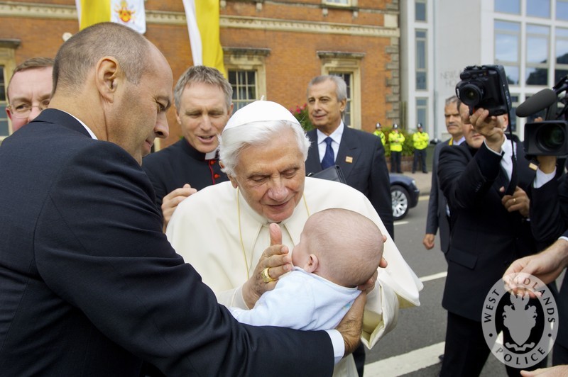 Pope Benedict greets a baby held by another man. There are guards in suits around them.