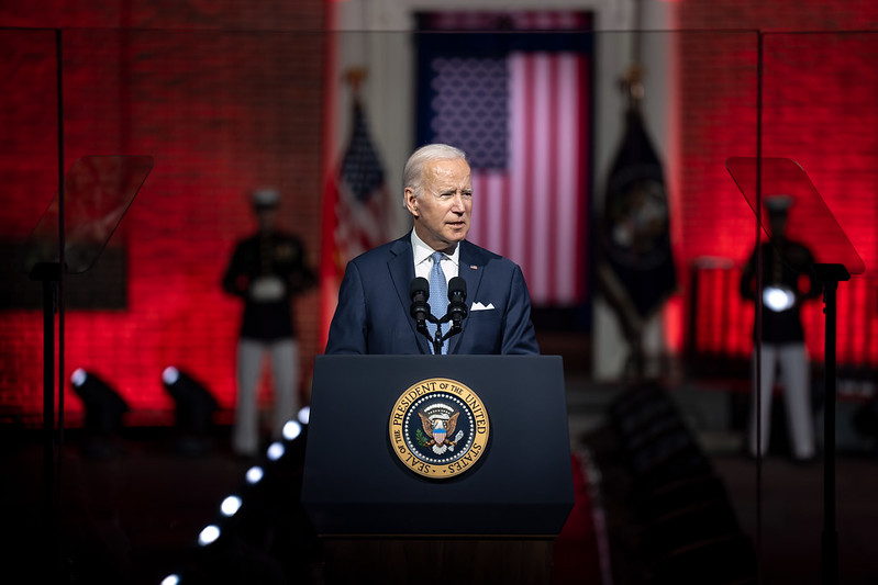 President Biden stands at a podium. Behind him is a large American flag. The lighting is deep red.