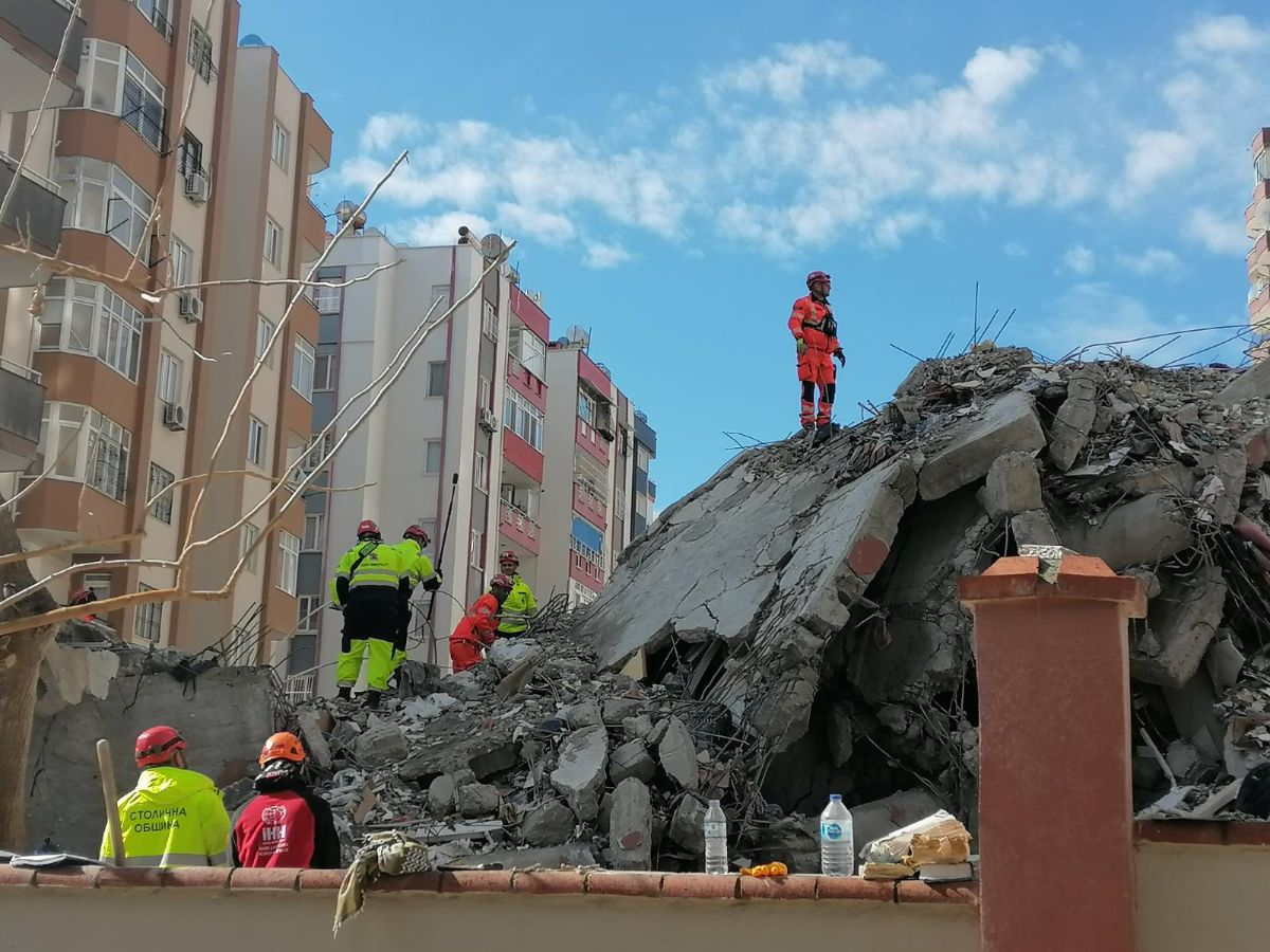 Several people in safety gear are assessing the pile from a collapsed building. The pile looks to be about 60 feet tall and about 100 feet wide when compared to the lone person standing on it.