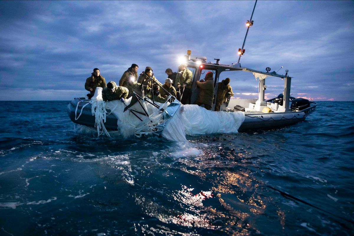A rigid hull inflatable boat carries a dozen sailors who are pulling the wreckage of a balloon out of the water