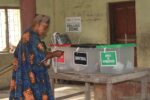 A single person in a room marked "polling zone" is placing their ballot into three tubs marked "presidential, senatorial, and house of representatives." The room looks older. The individual is wearing modern Nigerian clothing, a blue robe with bright orange spots and a hat