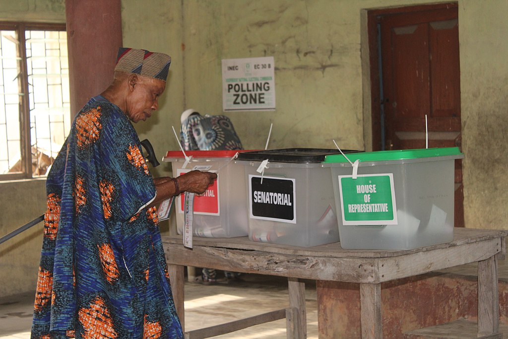 A single person in a room marked "polling zone" is placing their ballot into three tubs marked "presidential, senatorial, and house of representatives." The room looks older. The individual is wearing modern Nigerian clothing, a blue robe with bright orange spots and a hat