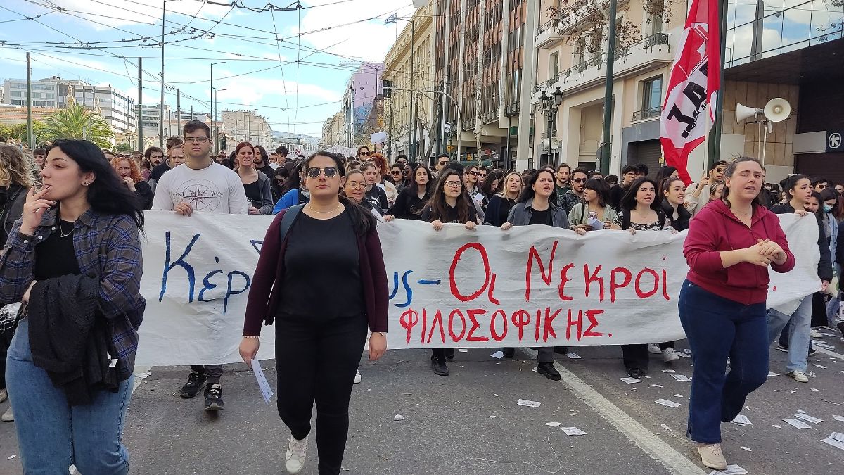 Dozens of people march a long a city street. They are carrying a banner with writing in Greek.