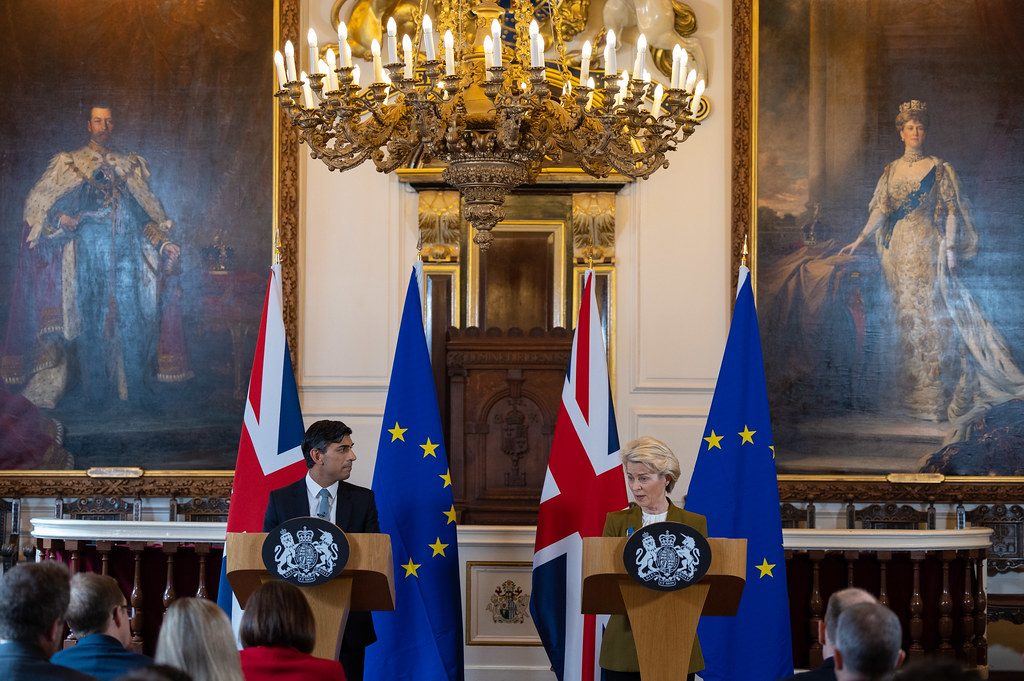 Two people in business suits stand at identical podiums in front of UK and EU flags in a grand room, under a large chandelier.