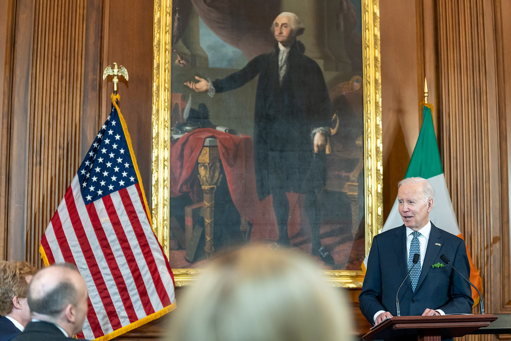 A large painted portrait of George Washington is in the center of this photo. On the very edge of the right portion of the photo you can see President Joe Biden standing in front of an Irish flag.