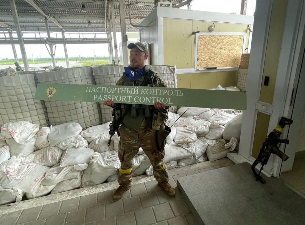 A man in military uniform-ish stands holding a border crossing sign written in Russian