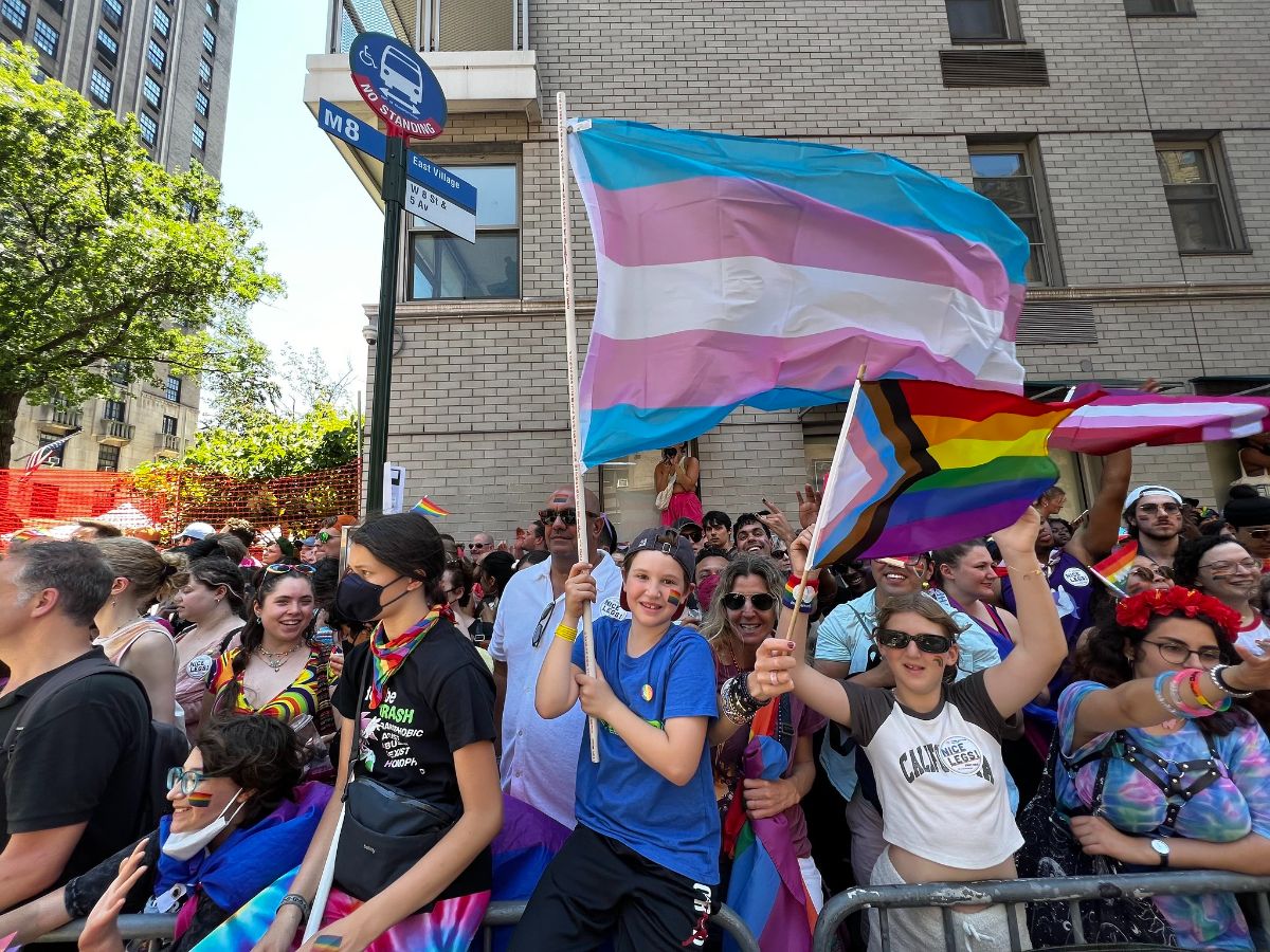 Hundreds of people are on a street corner, behind a safety fence. Three large pride flags (transgender pride, progress pride and lesbian pride) fly in the center of the group.