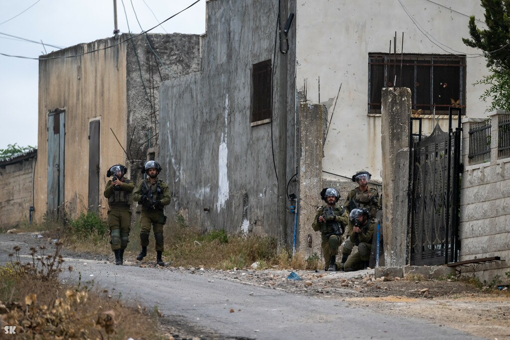 Two soldiers in patrol gear walk a street in front of rundown buildings. There are three more soldiers seeking cover behind a gate or fence.