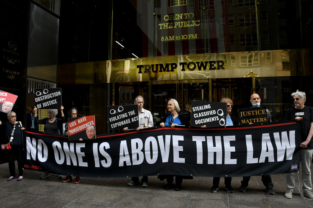 In front of the main entry to Trump Tower Manhattan about a dozen protestors stand holding a large banner that reads "No one is above the law"