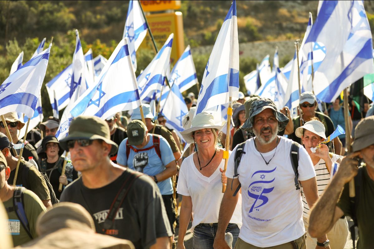 Dozens of people wearing hats to protect themselves from the sun march among a low hill. About 75% are carrying the flag of Israel.