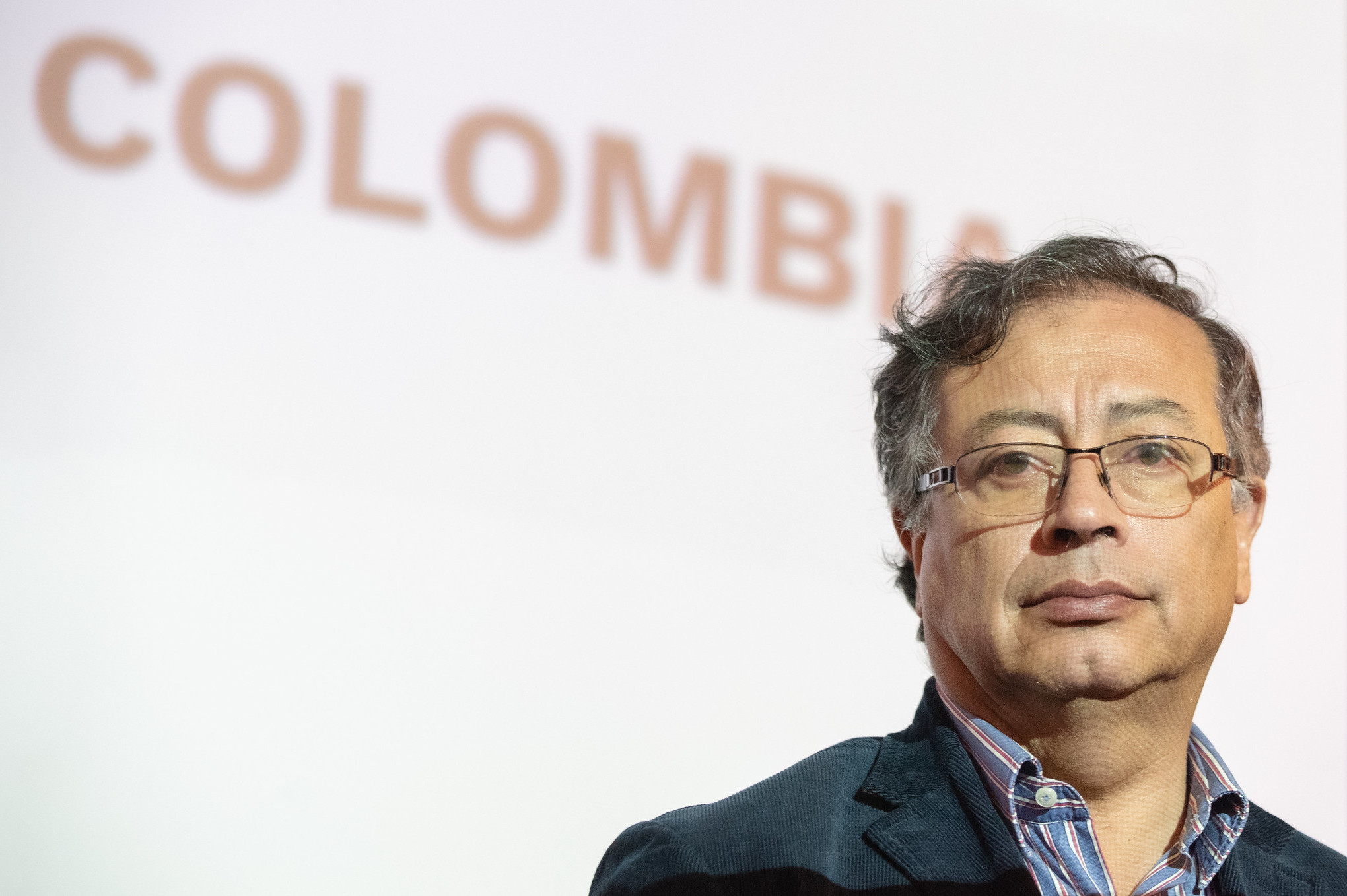 Gustavo Petro in front of the word "Colombia", wearing glasses and a button-up shirt and blazer.