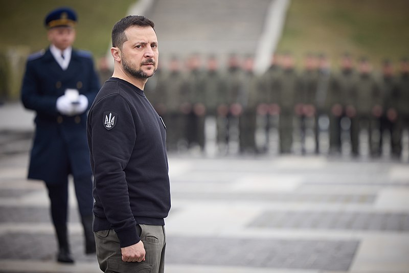 Volodymyr Zelenskyy stands in the foreground. Behind him is a man in well-kept official clothing. In the distance are hundreds of soldiers, out of focus.