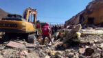 A large tracked digging machine sorts through heavy rubble as a single individual searches the remains of a building.
