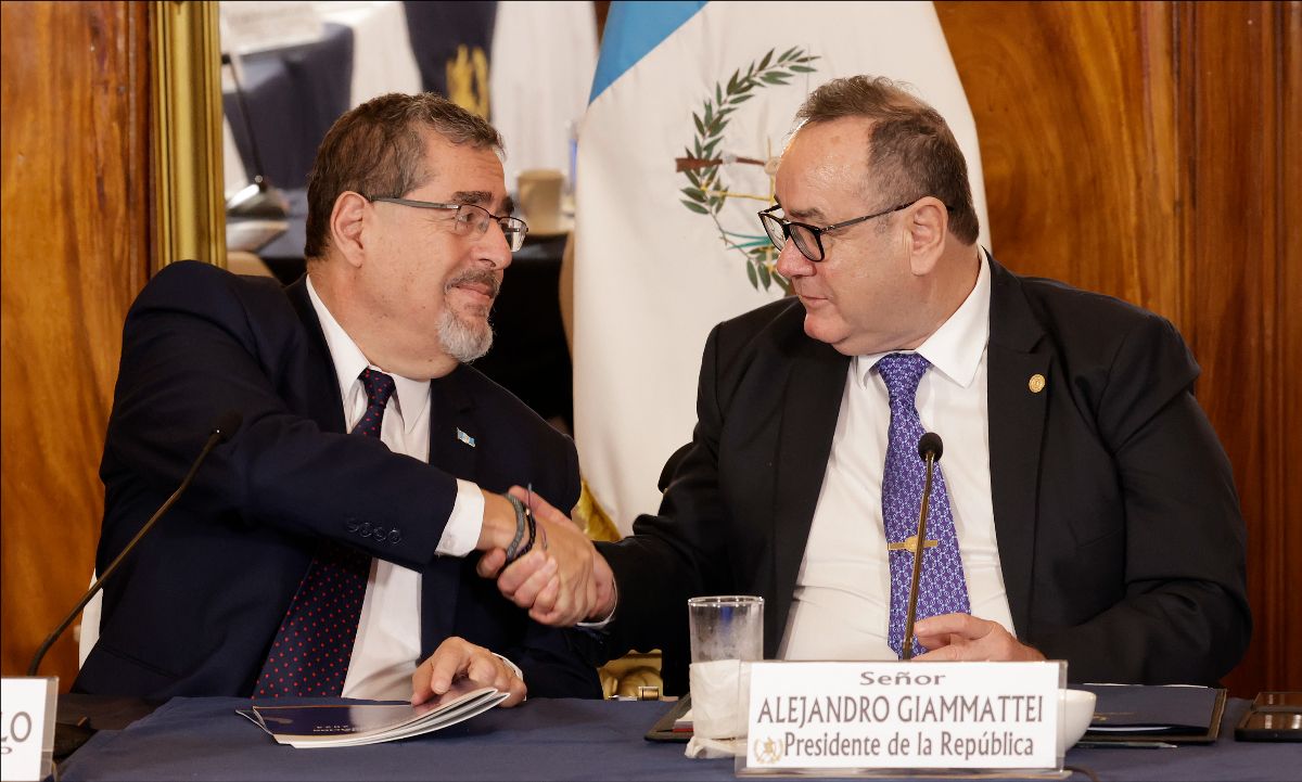 Two men shake hands while seated at a table in front of the flag of Guatemala.