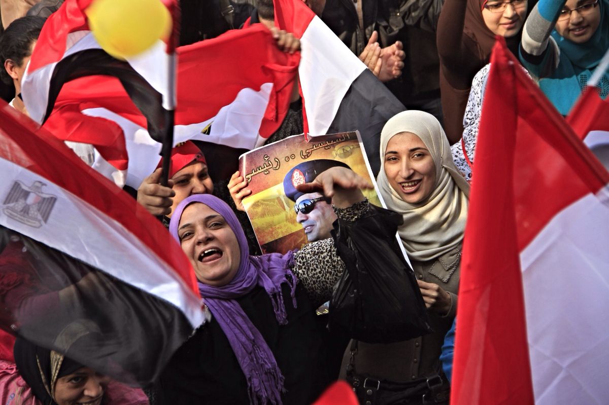 Faces from two women poke-out from a crowd waving flags. One holds a picture of General el-Sissi. The crowd is mostly smiling.