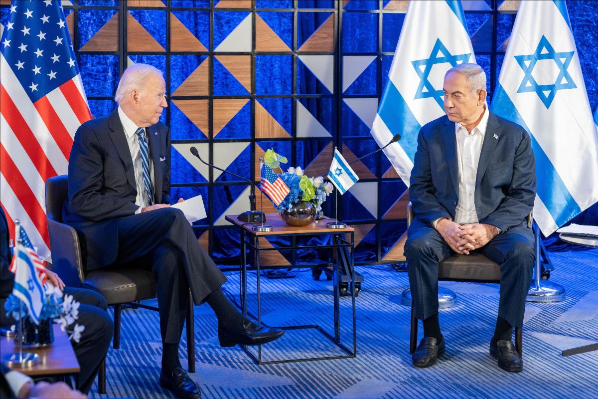 President Joe Biden sits across a side table from Prime Minister Benjamin Netanyahu. The flags of the United States and Israel are behind their respective leaders, with smaller flags on the table next to microphones pointed at each man.