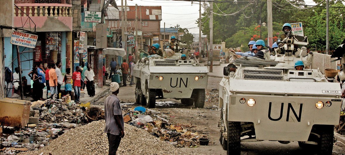 Two United Nations armored vehicles move through a street in Haiti. Many Haitians gather on the left side of the street watching the vehicles and UN blue hats drive by.