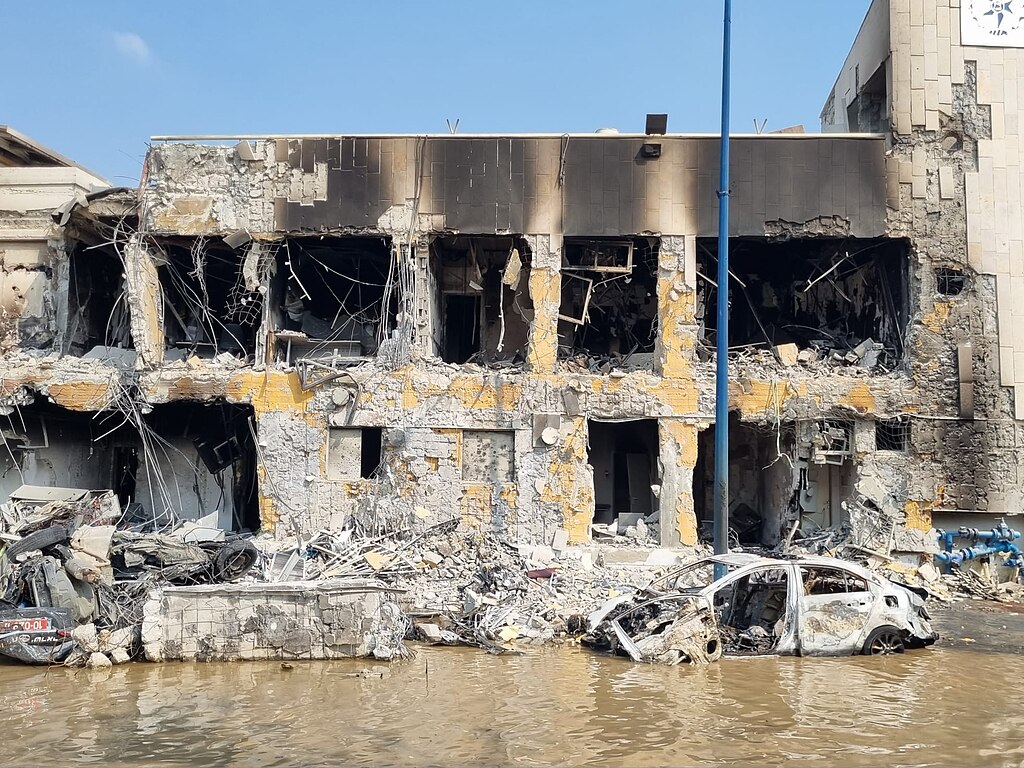 The shell of a two-story building with massive amounts of damage from fire and explosion. There is a burned out car in front of the building. The street is full of water.