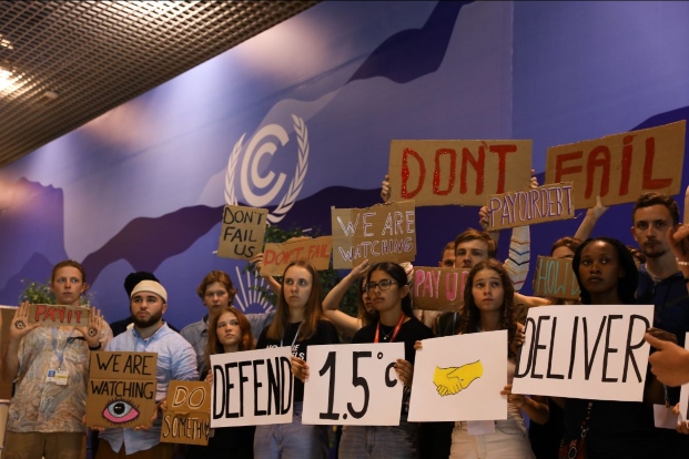 Several people hold signs calling for climate activism. The largest sign is a series that states "Defend 1.5 & deliver"