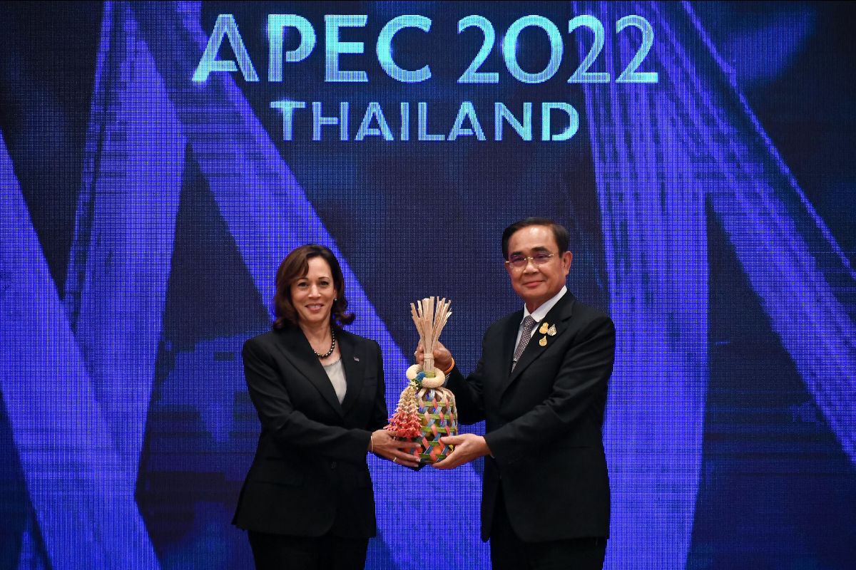Two people in suits hold an ornamental object in front of a sign that says APEC 2022 Thailand. There is a dark blue background with broad brighter blue stripes.