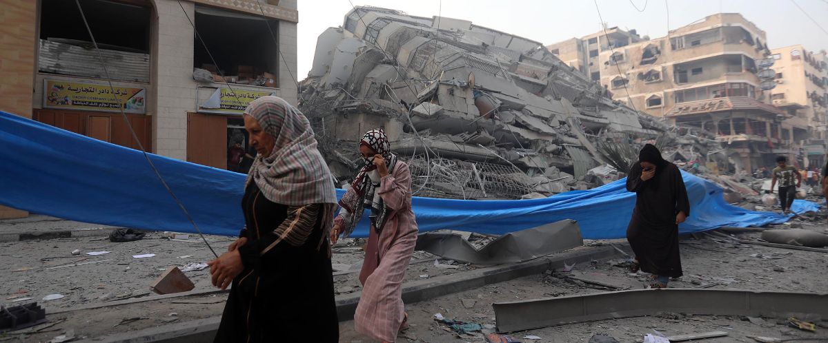 Three women with head coverings and long robes walk past a blue tarp that separates a road with some broken bits of a building. Behind the tarp is a collapsed multi-story building flanked by a two-story building with some damage and a five-story building with significant damage.