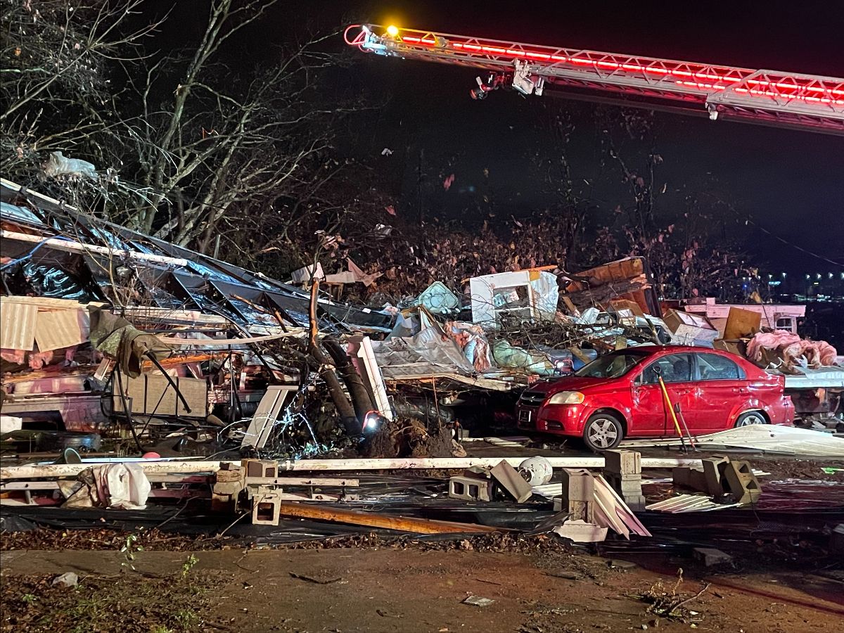 The boom of a firetruck rises over the wreckage of a house destroyed by a tornado. There is a red car with broken windows atop more wreckage. The tree to the left of the frame has roofing hanging inside of it.