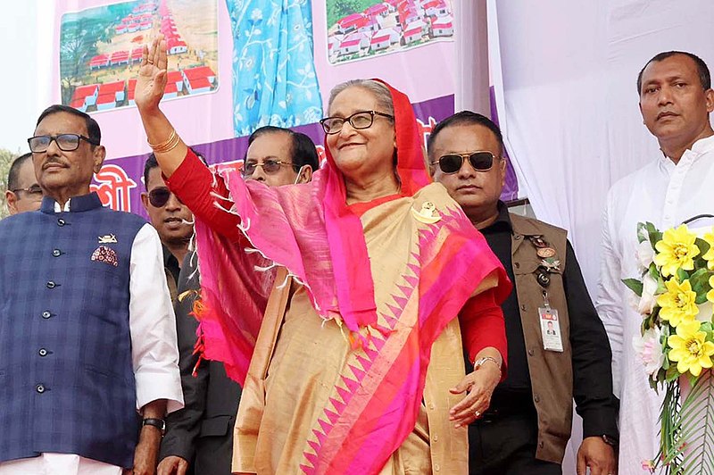 Woman with grey hair and glasses in a sari standing in front of four men in both Western and South Asian suits. Her sari is pink and yellow. She waves at a crowd.