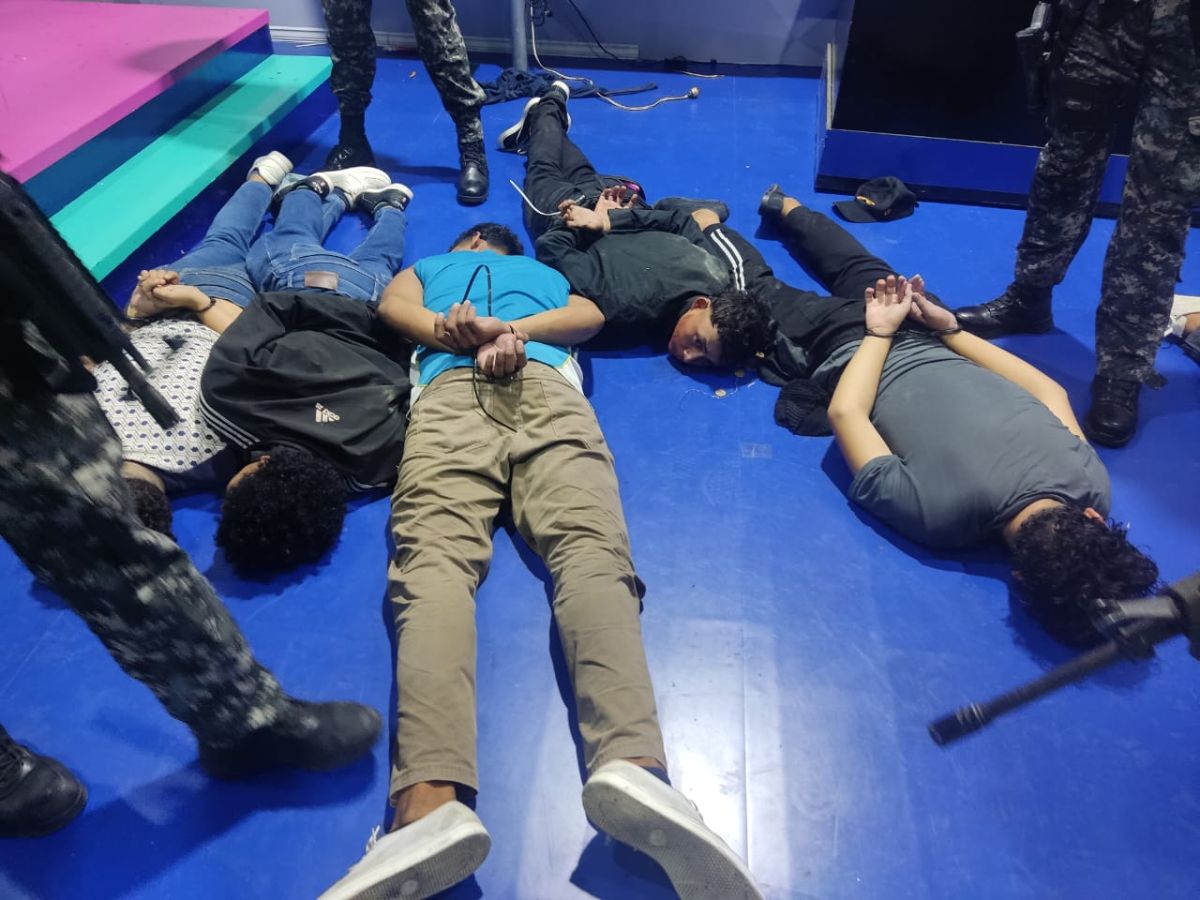 Several men lay face-down on the floor. Their hands are bound. They are dressed in t-shirts and jeans