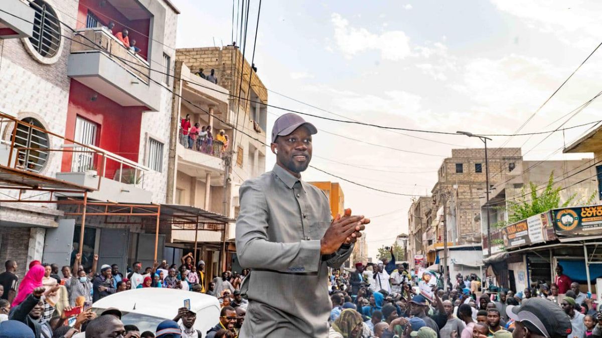 Senegalese opposition leader Ousmane Sonko stands on a raised platform in a grey suit with a matching cap. He is applauding. Surrounding him are the heads of a large crowd on a street as they watch his speech.