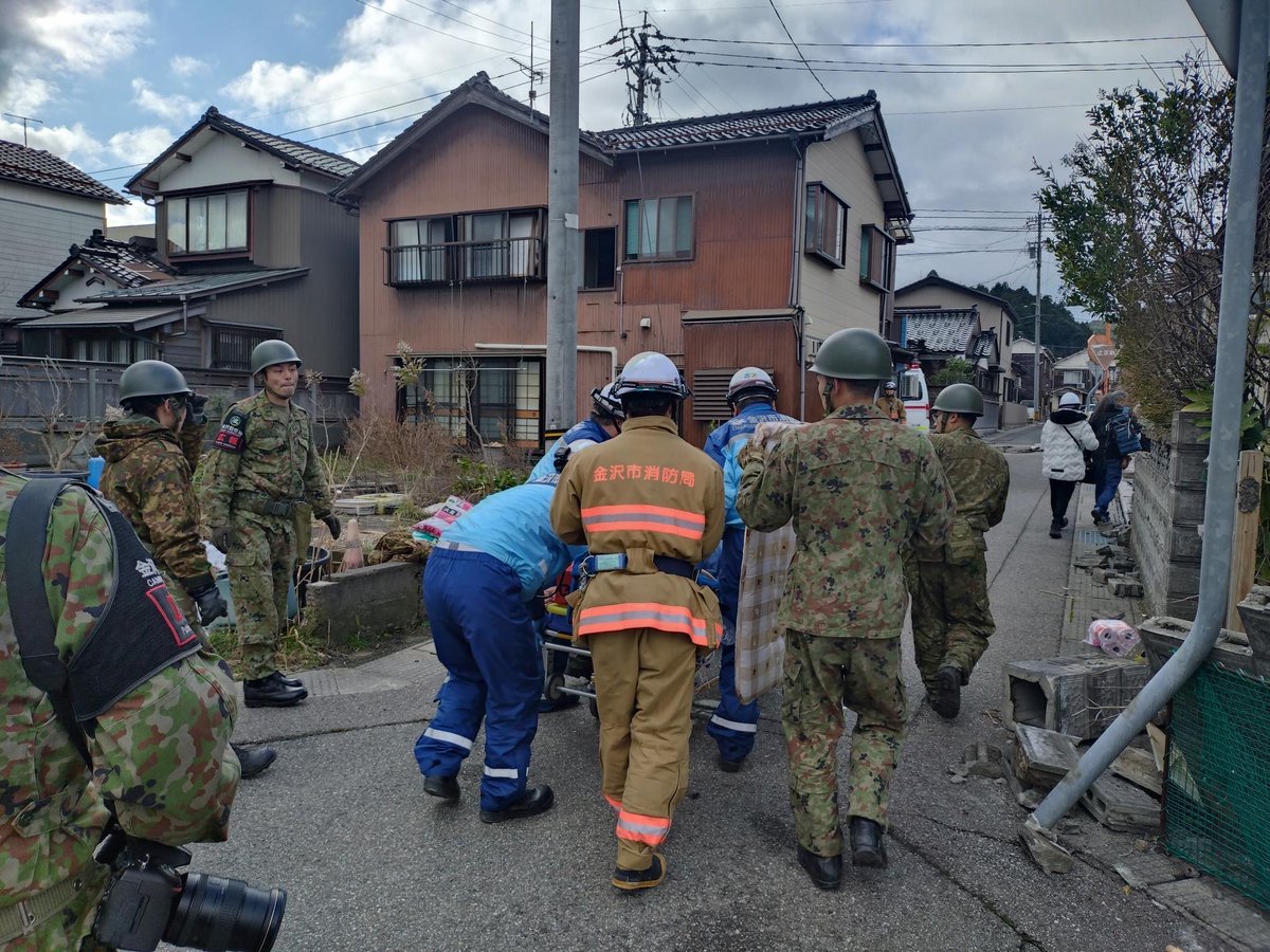 Soldiers and emergency workers push a cart through a small street. There may be a person on the cart, as one of the soldiers is holding up a blanket blocking view of what is there.  The houses show damage from an earthquake. 