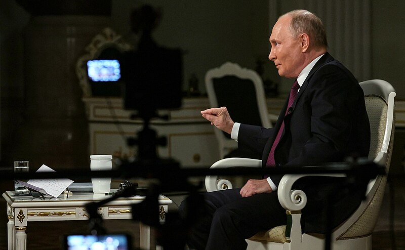 Sitting in a dining chair Russian President Vladimir Putin is facing to the left. There are cameras and video equipment around him.