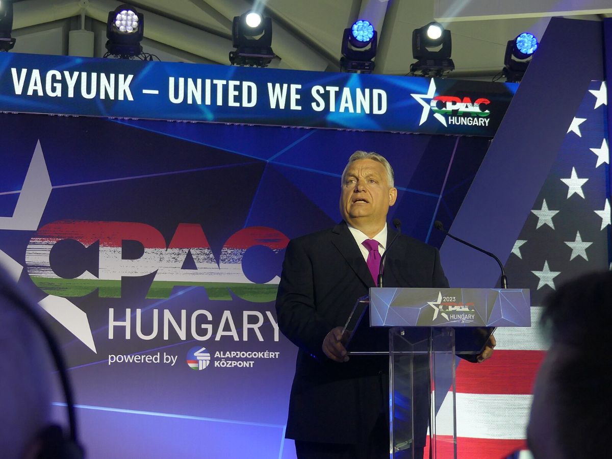 Standing slightly to the right is a man at a lectern. He is wearing a near-black suit with a red tie. Signage in English behind him says "United we stand" and "CPAC, Budapest Hungary."
