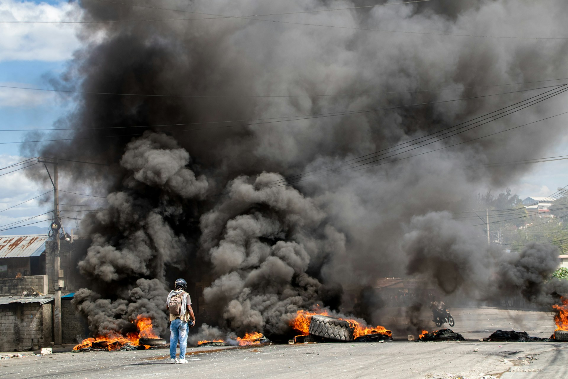 Lone human in jeans and tactical non-military gears stands with back to photographer. They are wearing a helmet. The person is looking at dark smoke burning from tire and other fires.
