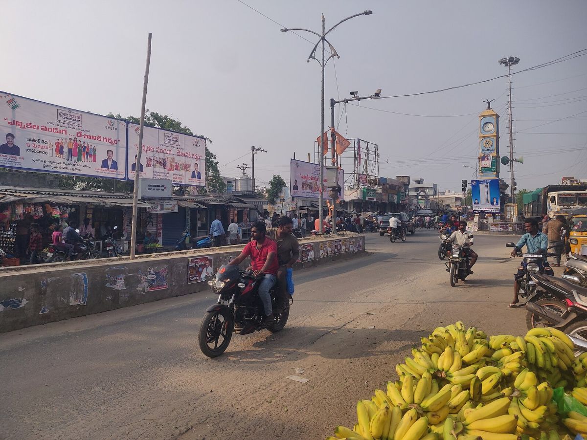 A man is riding a motorcycle down a busy street with a bunch of bananas on the side. The street is bustling with people and vehicles, with street lights illuminating the scene. The man is accompanied by a woman on the motorcycle. In the background, there are power lines and a poster visible. The image captures the vibrant street life in a city area. The colors in the image include shades of brown, gray, and yellow, creating a dynamic and lively atmosphere. This scene showcases a typical day in a bustling urban environment.