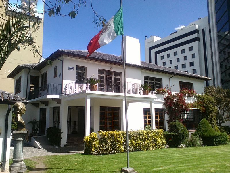 A large white building with a flag flying in the air is the focal point of this image. The flag is prominently displayed in the upper left corner, with a potted plant nearby. Another potted plant can be seen on a porch area to the right of the building. A few flowers are scattered throughout the scene, adding a pop of color. The building appears to be a hotel or a luxury residence, with a well-maintained lawn in front of it. The overall aesthetic is clean and elegant, with a touch of natural elements enhancing the ambiance.