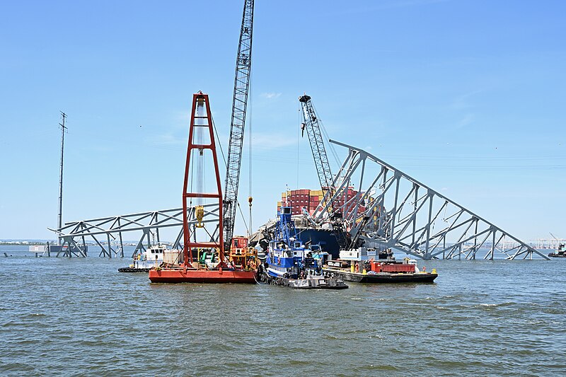 Maritime construction scene with barges and cranes working on an incomplete steel girder structure, with a container ship in the background under a clear blue sky.