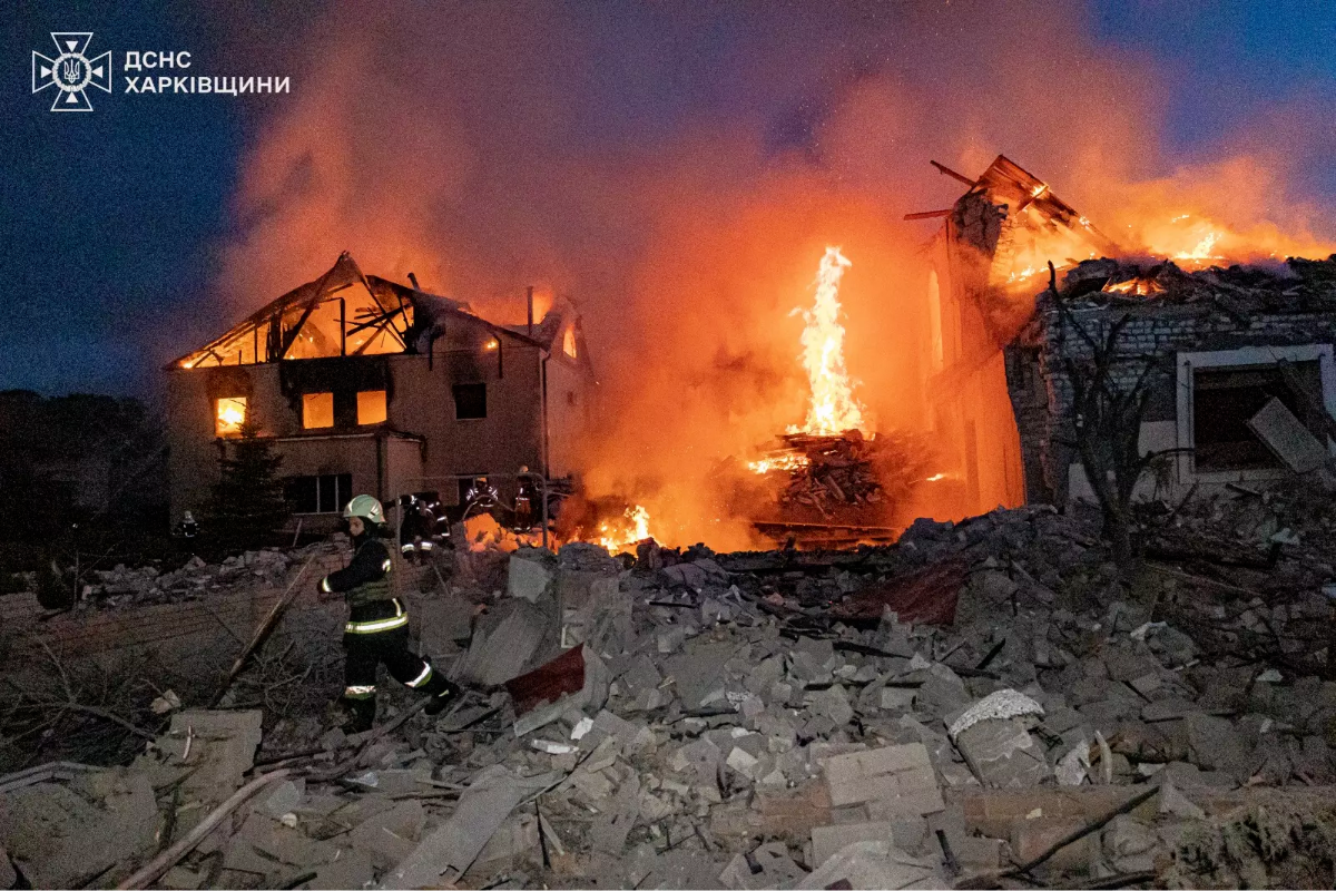 A firefighter moves amidst rubble while a large building burns ferociously at night, with flames and smoke filling the scene.

Text Presented in the Image: The top left corner of the image contains Cyrillic text "ДСНС ХАРКІВЩИНИ" along with an emblem, indicating the image relates to emergency services in the Kharkiv area.