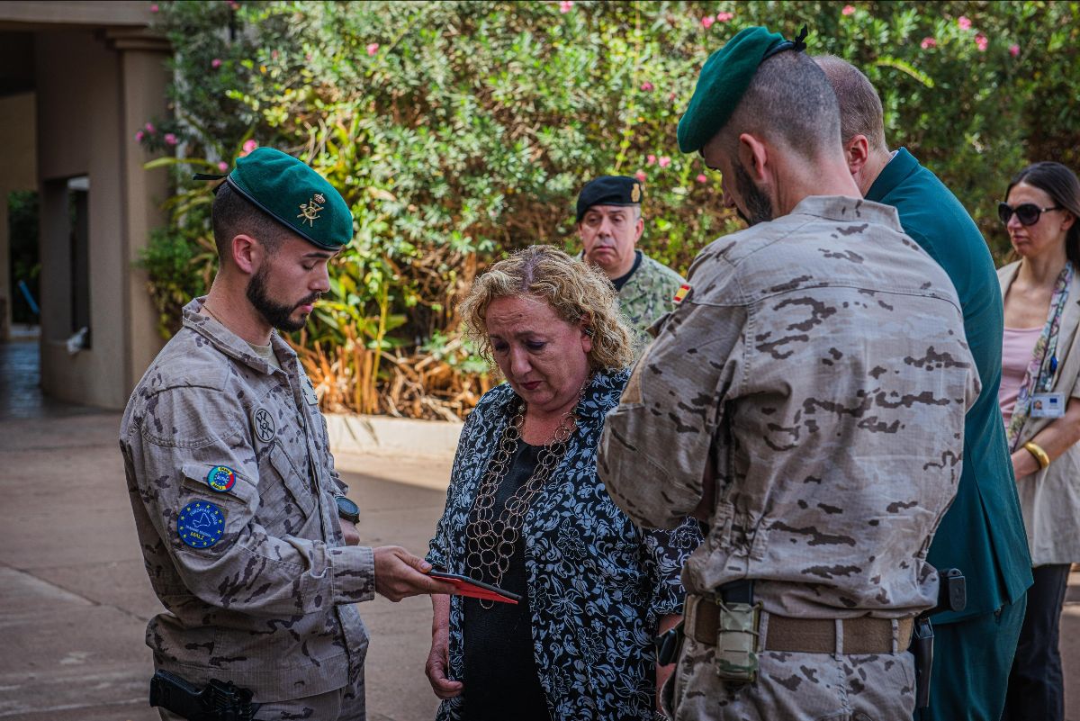 Two soldiers in desert camouflage and green berets present a document to a civilian woman in a patterned top, with onlookers in the background against a backdrop of greenery and flowers.