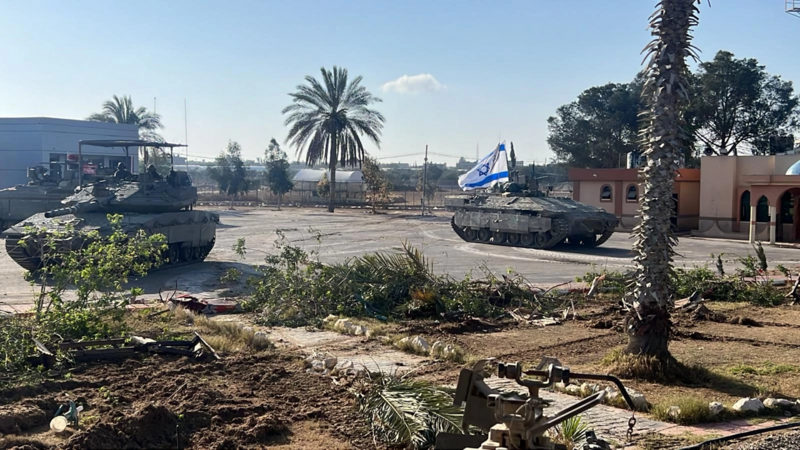 Two camouflaged tanks with an Israeli flag display, staged on pavement against a backdrop of palm trees and a clear blue sky.