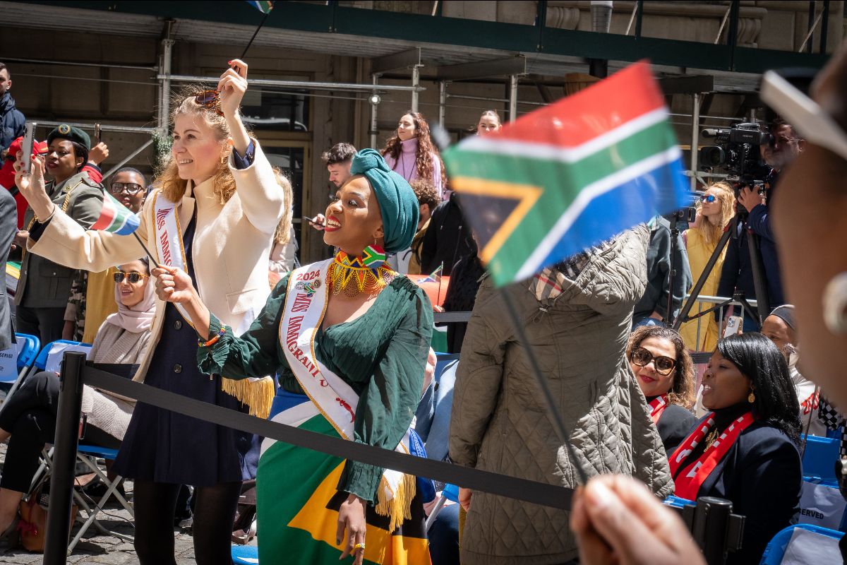 People waving South African flags at an outdoor event, with a woman wearing a sash that reads “2024 MISS IMMIGRANT USA.”