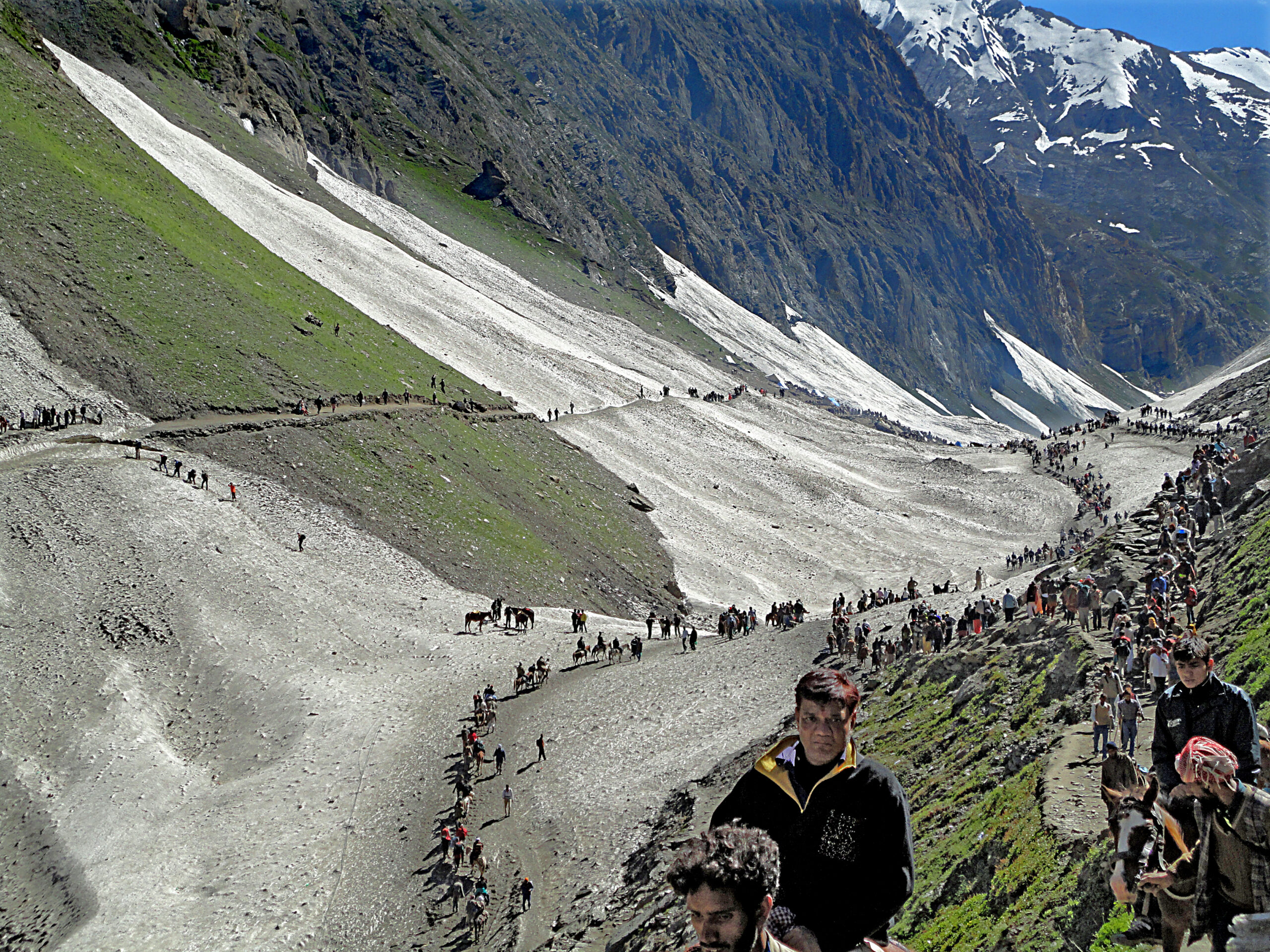 A busy mountainous path with numerous people walking and on horseback amidst snowy and green slopes.