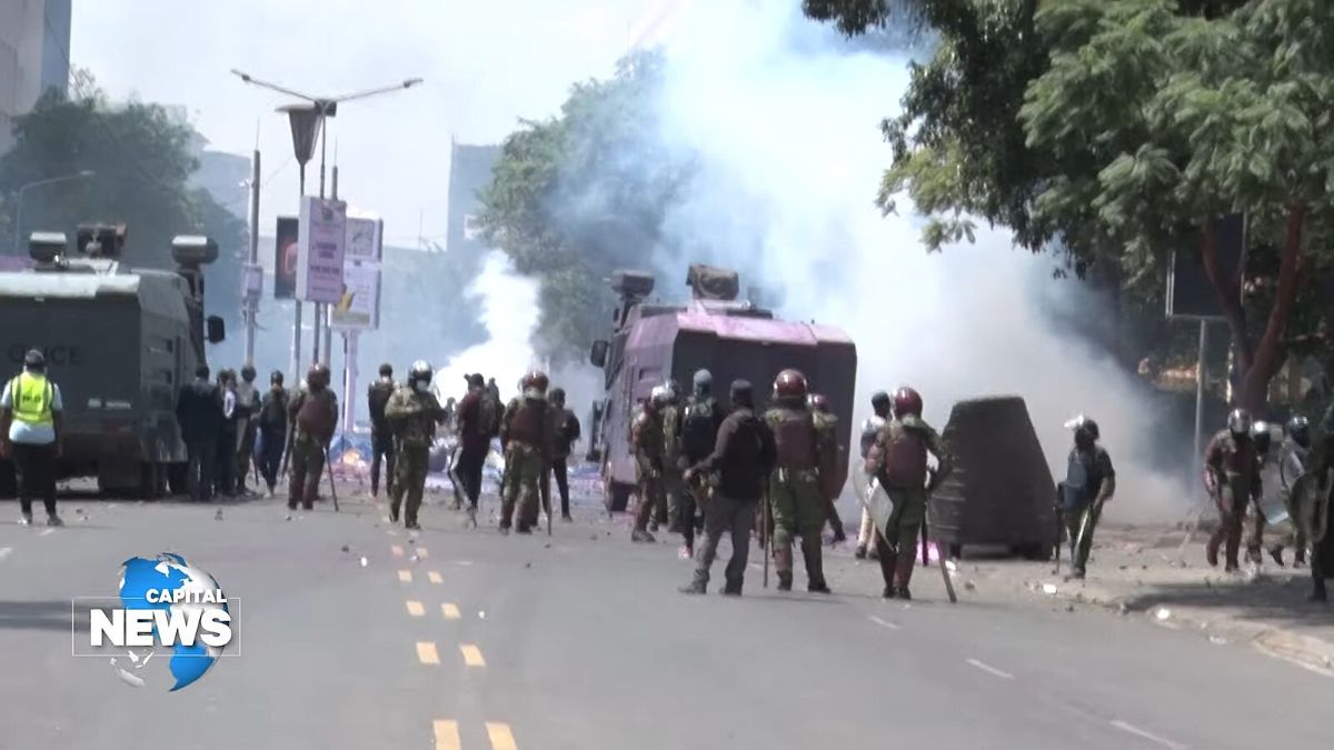 Street with uniformed police officers in protective gear, armored vehicles, and smoke in the background.