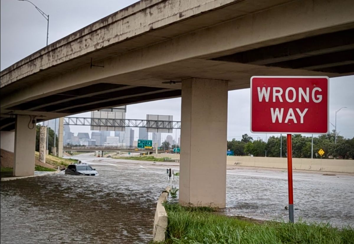 Flooded road under an overpass with a "WRONG WAY" sign and a partially submerged car.