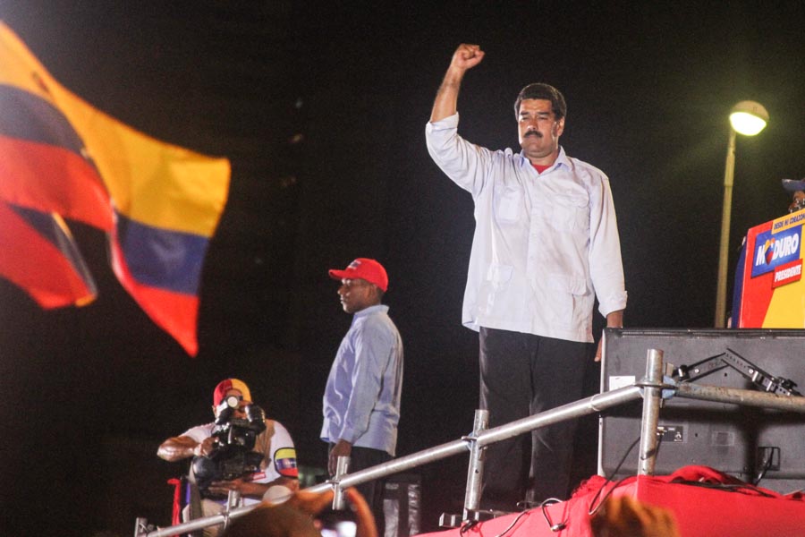 A man on an elevated platform raising his fist, with a large flag in the foreground and another man in a red cap nearby.
