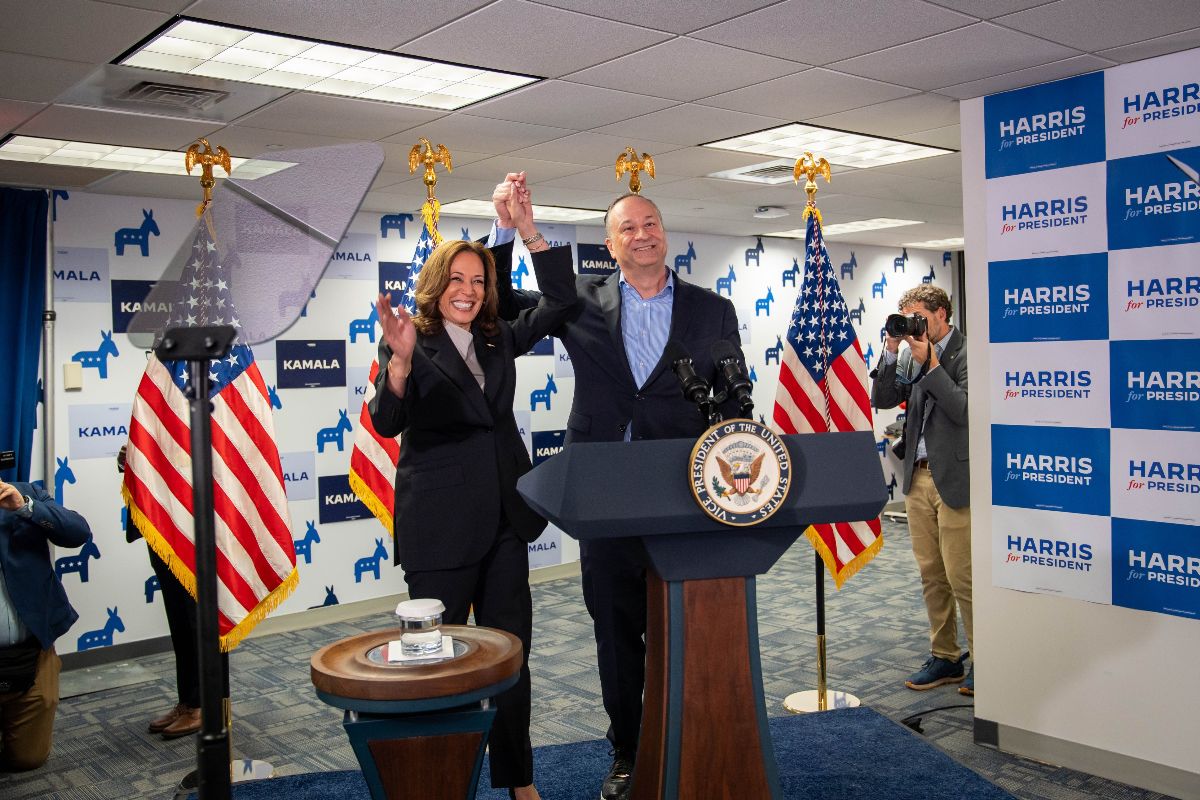 Man and woman celebrating on stage with hands raised, standing behind the Vice President's lectern, American flags, and campaign signs in the background.