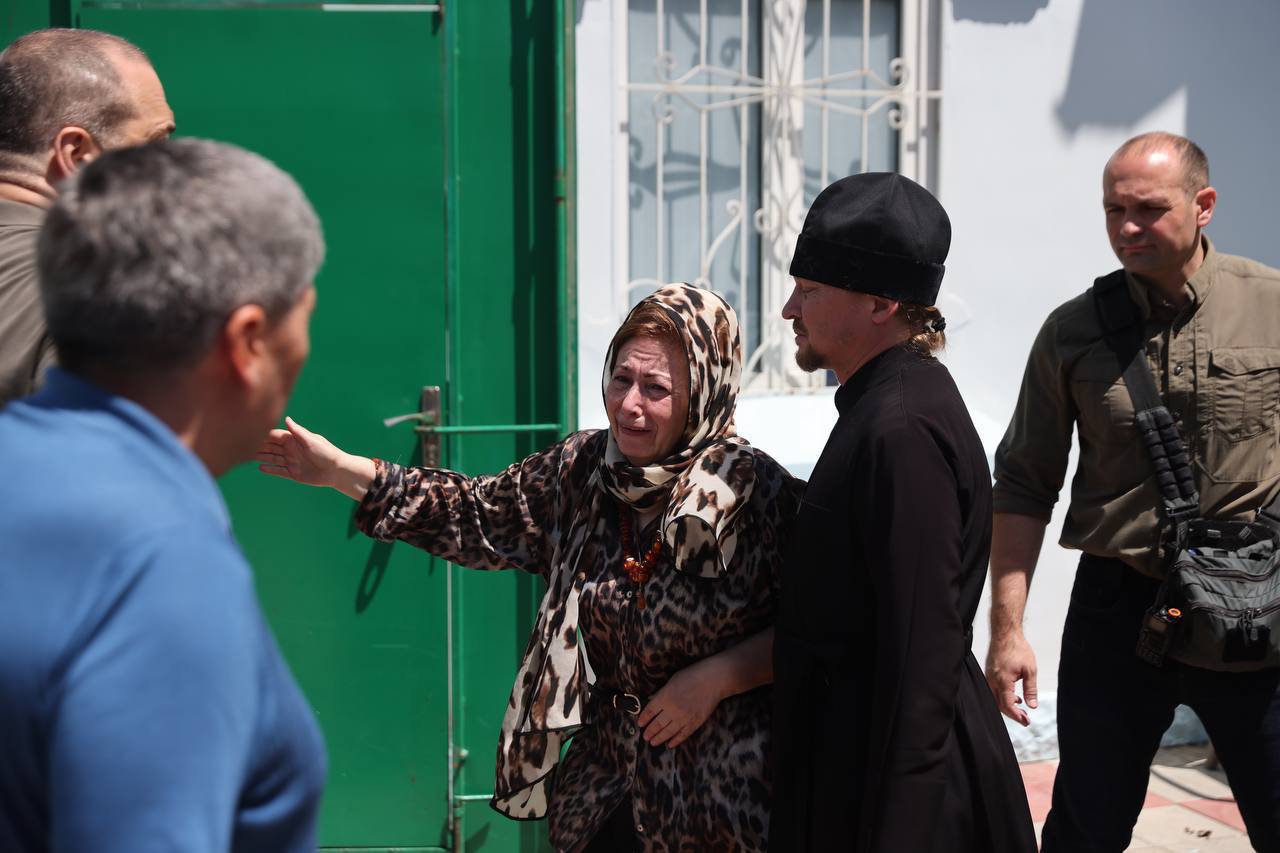 A distressed woman in a leopard-print headscarf gestures towards a man in a black robe.