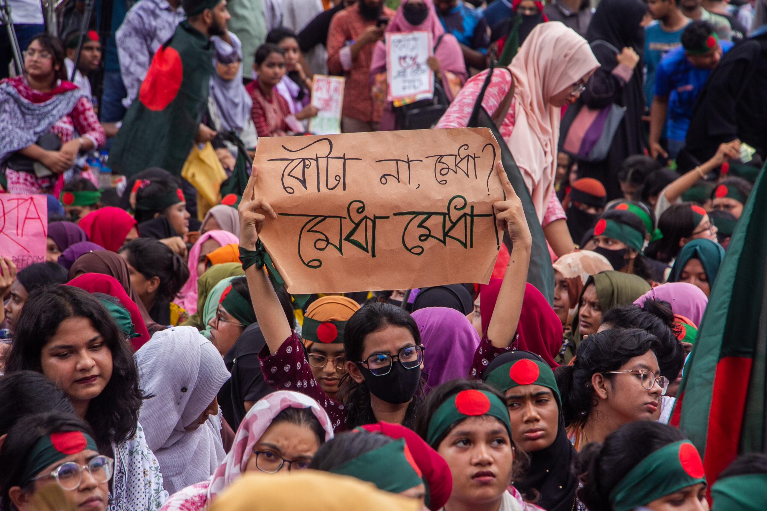 A large group of protesters, mostly women, with one holding up a brown sign with Bengali text.